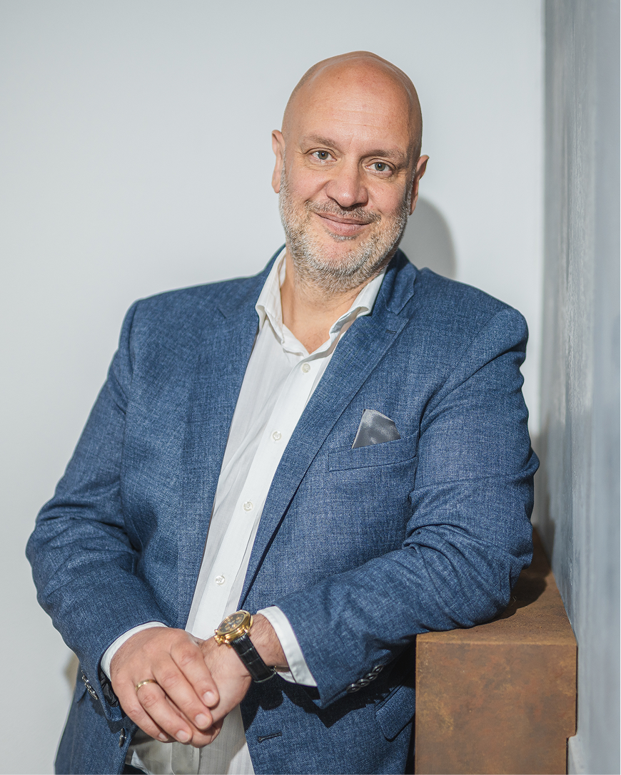 Smiling bald man in a blue blazer and white shirt leaning against a light wall, hands clasped over a wooden ledge.